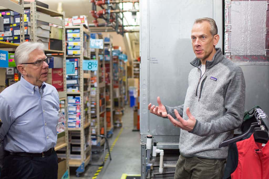 Two men standing in the stock room of a shoe store discussing the energy efficiency of their HVAC system.